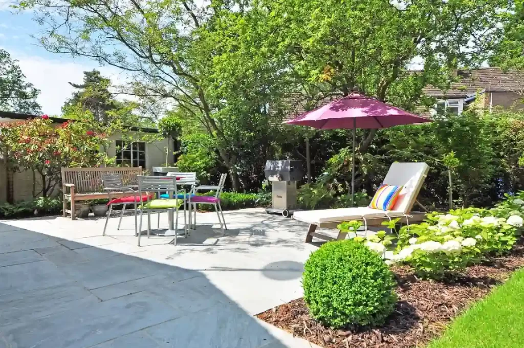 Uma área externa bem decorada com móveis ao ar livre, sombra de guarda-sol rosa, árvores verdes e plantas, ideal para relaxar e receber visitas com estilo e funcionalidade.