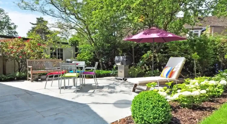 Uma área externa bem decorada com móveis ao ar livre, sombra de guarda-sol rosa, árvores verdes e plantas, ideal para relaxar e receber visitas com estilo e funcionalidade.
